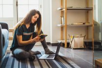 Girl sitting on rug and watching notebook while using laptop. — Stock Photo