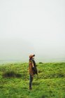 Side view of woman in warm jacket standing and looking up in foggy green grasslands. — Stock Photo