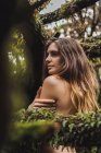 Naked woman embracing shoulder among mossy tree branches and looking away. — Stock Photo
