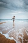 Junge Frau in Kleid, die im flachen Wasser der Ozeanwelle unter düsterem Himmel wandelt. — Stockfoto