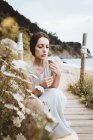 Young serious brunette in long dress posing on wooden beach pathway with dry flower in hands. — Stock Photo