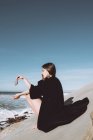 Elegante morena de larga chaqueta negra sentada en la roca y sosteniendo piedra pequeña con el océano en el fondo . - foto de stock
