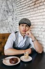 Thoughtful man in vintage cap sitting at table with coffee and donuts in cafe. — Stock Photo