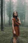 Confident young brunette in short dress posing in foggy dark woods and looking at camera. — Stock Photo
