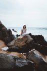 Mulher morena em vestido longo sentado na rocha molhada na costa do oceano . — Fotografia de Stock