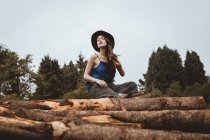 De baixo tiro de mulher bonita em roupa elegante sentado em troncos com árvores no fundo . — Fotografia de Stock