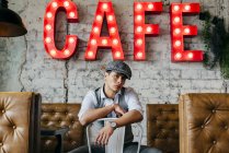 Hombre en ropa vintage posando en la silla en la cafetería y mirando a la cámara - foto de stock