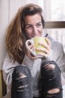 Cheerful woman sitting at window and relaxing with cup at home. — Stock Photo
