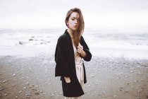 Стильная девушка в модной черной куртке, неэмоционально смотрящая на камеру на фоне океана в движении . — стоковое фото