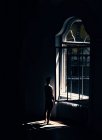 Vista lateral do homem irreconhecível em pé na janela no quarto escuro em casa . — Fotografia de Stock