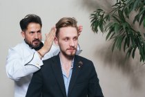 Barber styling hair of young man in suit in hairdressing salon. — Stock Photo