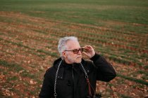 Elderly serious man in sunglasses and black jacket looking away with confidence on background of field. — Stock Photo