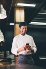 Усміхнений кухар стоїть на кухні ресторану і використовує смартфон . — стокове фото