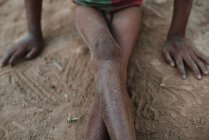 Crop dirty legs of ethnic kid sitting on sand. — Stock Photo