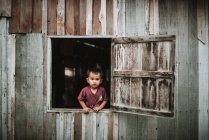 LAOS, 4000 ISLAS ÁREA: Adorable niño en ropa casual mirando por la ventana de la casa de madera del pueblo . - foto de stock