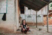 NONG KHIAW, LAOS: Mulher asiática descalça sentada perto de casa na rua da aldeia e olhando para a câmera . — Fotografia de Stock