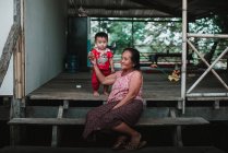 LAOS, 4000 ISLAS ÁREA: Mujer mayor sentada en las escaleras de la casa de madera y cogida de la mano del niño . - foto de stock