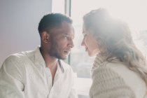 Couple in sweaters bonding by window and looking at each other — Stock Photo