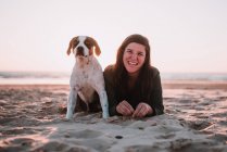 Porträt einer fröhlichen Frau mit kleinem Hund, der auf Sand liegt und in die Kamera blickt — Stockfoto