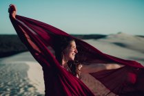 Vista lateral de mujer alegre en tela roja posando en dunas de arena - foto de stock