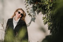 Fröhliche Frau in warmer Kleidung und Sonnenbrille an Wand und Busch. — Stockfoto