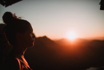 Vista lateral mulher atraente em pé no fundo do pôr do sol nas montanhas . — Fotografia de Stock