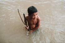 LAOS, 4000 ÎLES : Garçon avec des bâtons debout dans une rivière sale — Photo de stock