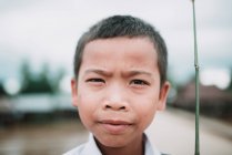 Laos, 4000 Inseln: ernster Junge blickt auf verschwommenem Dorfhintergrund in die Kamera. — Stockfoto
