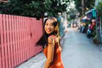 Cheerful woman in red dress posing on street and looking over shoulder at camera — Stock Photo