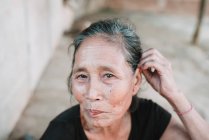 NONG KHIAW, LAOS: Elderly woman adjusting hair while sitting on village street. — Stock Photo
