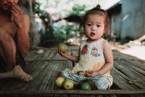 NONG KHIAW, LAOS: Chica bonita sentada en la puerta de madera en la aldea y sosteniendo fruta . - foto de stock