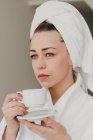 Задумчивая женщина в полотенце стоя с керамической чашкой и глядя в сторону . — стоковое фото