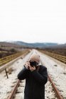 Fotógrafo irreconhecível em pé na estrada de ferro vazia e apontando com câmera . — Fotografia de Stock