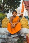 LAOS- FEBRUARY 18, 2018: Little  monk boy in orange clothes looking at camera and sitting on fence. — Stock Photo