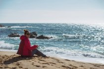 Зрелая женщина сидит на берегу моря в солнечный день и смотрит в камеру — стоковое фото