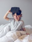 Sonriente chico acostado en la cama y quitándose gafas VR - foto de stock