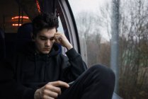 Hombre reflexivo sentado y utilizando el teléfono inteligente en el autobús en el día lluvioso
. - foto de stock