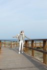Cheerful young stylish woman standing with hands apart on boardwalk at seaside. — Stock Photo