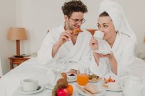 Alegre pareja joven en albornoces sentado y desayunando en la habitación del hotel . - foto de stock