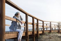 Mujer joven soñadora sentada en una pasarela de madera y mirando hacia otro lado . - foto de stock