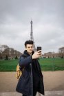 Giovane uomo in piedi con il telefono e prendendo selfie sullo sfondo della torre Eiffel . — Foto stock