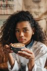 Curly mulher bebendo xícara de café e olhando para a câmera — Fotografia de Stock