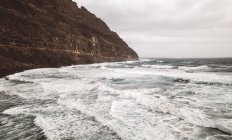 View to dark coastal cliff and wavy stormy ocean in cloudy day. — Stock Photo