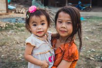 LAOS- FEBRERO 18, 2018: Hermanas jóvenes y alegres de pie en la aldea y mirando a la cámara . - foto de stock