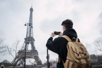 Vista posteriore del giovane in piedi con il telefono e scattare foto della torre Eiffel . — Foto stock
