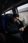 Nachdenklicher junger Mann sitzt bei Regen im Bus und benutzt Smartphone. — Stockfoto