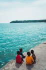 Vista posterior de niños étnicos sentados en terraplén de hormigón en el océano azul . - foto de stock