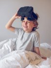 Mignon garçon couché sur le lit et enlever les lunettes VR — Photo de stock
