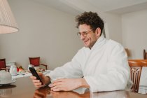 Sorrindo homem em roupão de banho navegando smartphone no quarto de hotel — Fotografia de Stock