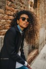 Stylish curly woman in sunglasses at brick wall — Stock Photo